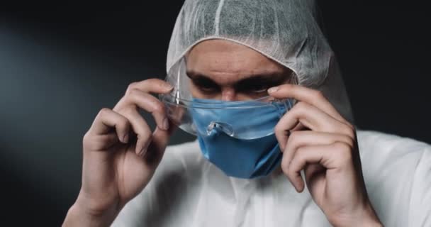 Egy fiatal, fáradt, fehér férfi orvos teljes védelemben, aki leveszi a szemüvegét és a kamerába néz. Portré epidemiológusról, aki feldúltan és elégedetlenül néz le. Coronavirus koncepció. Kovid-19 — Stock videók