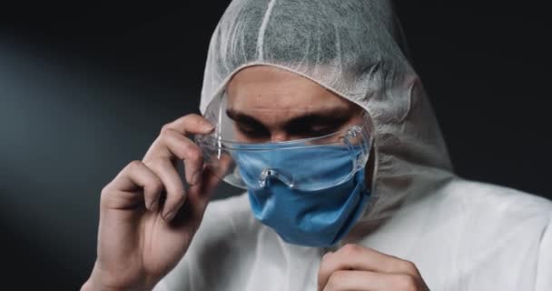 Крупным планом уставшего молодого кавказского эпидемиолога-мужчины в защитных очках и медицинской маске. Портрет мужчины-врача, смотрящего в камеру расстроенным. Концепция коронавируса. Медик из Ковид-19 . — стоковое видео