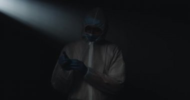 Beyaz erkek doktor epidemiyolojistin portresi. Tıbbi maskeli ve gözlüklü. Plastik eldiven takıyor ve siyah arka plandaki kameraya bakıyor. Covid-19 salgını.