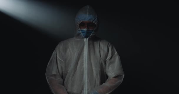 Портрет молодого кавказского врача-эпидемиолога в защитном костюме с медицинской маской и очками, смотрящего в камеру усталыми глазами на черном фоне. Медик во время эпидемии коронавируса — стоковое видео