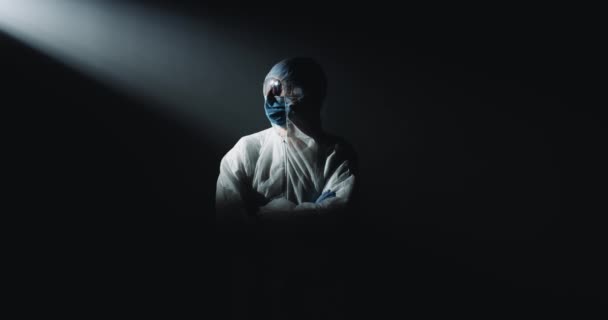 Portrét plně vybaveného zdravotníka v bílém sterilním kostýmu, brýlích a lékařské masce, stojícího ve tmě před kamerou. Doktor v ochraně před pandemií covid-19. — Stock video