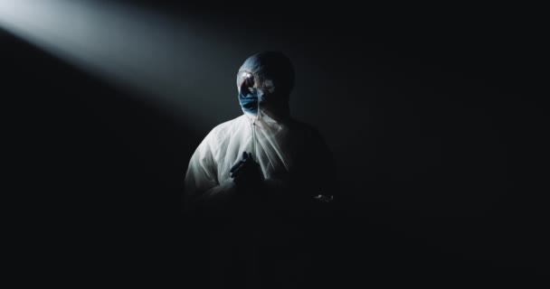 Portret w pełni wyposażonego epidemiologa w białym sterylnym kostiumie, goglach i masce medycznej stojącej w ciemności przed kamerą. Lekarz w ochronie przed pandemią koronawirusa. — Wideo stockowe