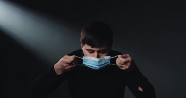Mavi tıbbi maske takan beyaz bir gencin portresi ve arka planda ciddi üzgün bir suratı olan kameraya bakıyor. Coronavirus salgını. Güvenlik kavramı.