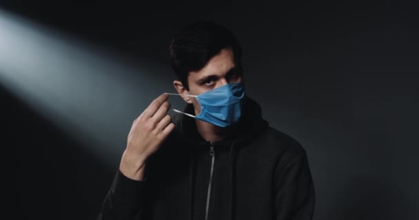 Porträtt av vit ung stilig man som tar på sig medicinsk mask och tittar på kameran med sorgset ansikte. Mörk vägg bakgrund. — Stockvideo