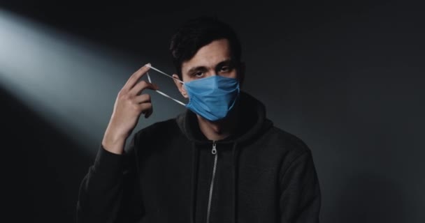 Portrait of Caucasian young sad male taking on blue medical mask and looking at camera with upset face. Black wall background. Guy during coronavirus outbreak. — Stock Video