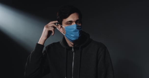 Портрет белого молодого расстроенного парня, надевающего синюю медицинскую маску и смотрящего в камеру с грустным лицом. Фон черной стены. Пациент во время пандемии коронавируса . — стоковое видео