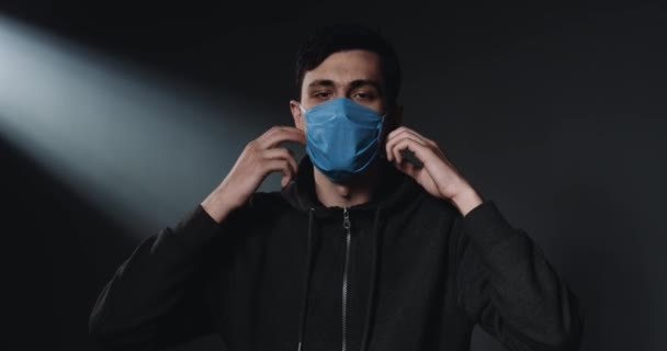 Portrait shot of Caucasian young handsome man with moustache taking off medical mask and looking at camera with sad face. Dark wall background. Covid-19 virus protection. — Stock Video