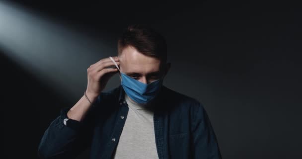 Beyaz, üzgün, genç bir erkek hastanın portresi mavi tıbbi maske takıyor ve üzgün bir yüzle kameraya bakıyor. Siyah duvar arkaplanı. Koronavirüs salgını sırasındaki adam. Covid-19 konsepti. — Stok video