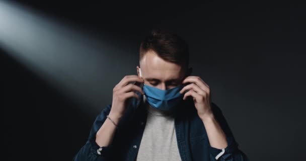 Retrato de un hombre caucásico joven guapo quitándose la máscara médica azul, su protección contra el coronavirus. Fondo oscuro de la pared. Concepto pandémico de Covid-19. Feliz chico alegre sonriendo a la cámara . — Vídeo de stock