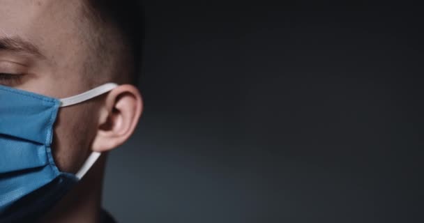 Close up of half face of Caucasian young guy in blue medical mask turning eyes and looking at camera. Portrait of male patient on black wall background. Covid-19 concept. — Stock Video