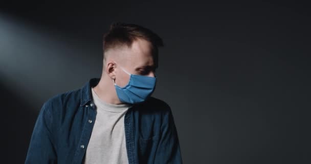 Caucasian young handsome man in medical blue mask caughing and sneezing, covering face with arm and elbow on black wall background. Coronavirus symptoms concept. Male patient with flu or cold. — Stock Video