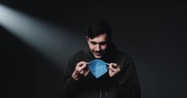 Portret van een blanke jonge gelukkige mannelijke patiënt die een blauw medisch masker afdoet en glimlacht naar de camera met een vrolijk gezicht. Zwarte muur achtergrond. Guy tijdens uitbraak van het coronavirus. — Stockvideo