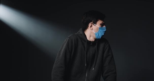 Paciente do sexo masculino, branco, com máscara azul médica, tosse e espirros, cobrindo o rosto com o braço e o cotovelo no fundo da parede escura. Conceito de sintomas de coronavírus. Homem com gripe ou frio . — Vídeo de Stock