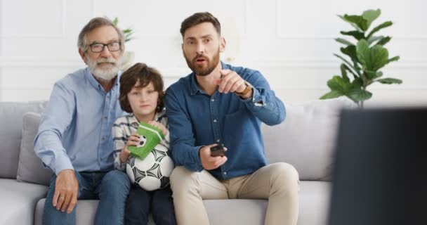 Caucasian male three generations. Little cute kid with old grandfather and father sitting on couch in living room and watching sport channel with football on TV. Senior man with son and grandson. — Stock Video