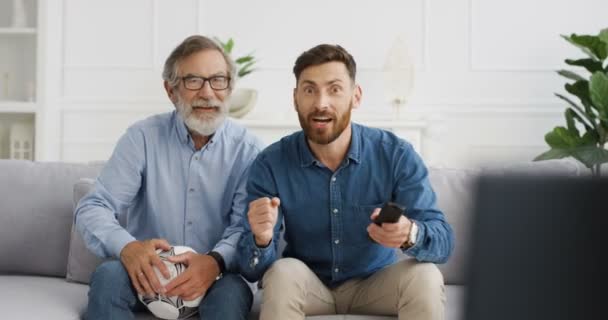 Vit man i två generationer. Gråhårig gammal far och vuxen son sitter på soffan i vardagsrummet och tittar på sport kanal med fotboll på TV. Senior och unga män uppmuntrande upp för sport match. — Stockvideo