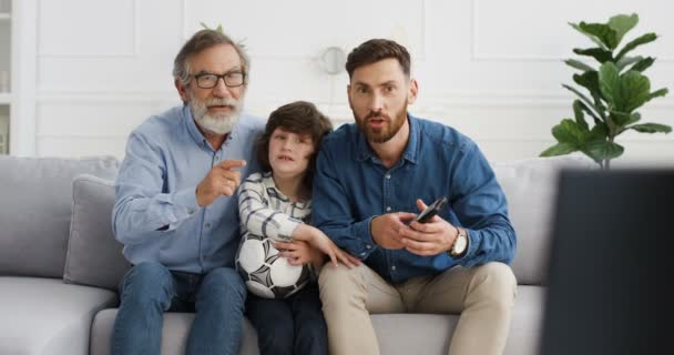 Kavkazská rodina, tři generace. Malé dítě se starým dědečkem a otcem sedí doma na pohovce a dívá se na sportovní kanál s fotbalem v televizi. Starší muž se synem a vnukem odpočívající uvnitř. — Stock video