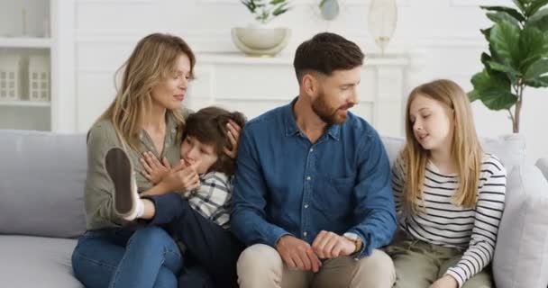 Portrét laciné bělošské rodiny se dvěma dětmi sedícími na gauči a radostně se usmívajícími při objímání a mluvení. Malý roztomilý chlapec a dívka s matkou a otcem doma. Rodiče s malými dětmi — Stock video