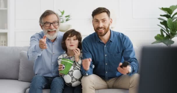 Caucasian males generations. Small child with old grandfather and father sitting on sofa at home and cheering up for goal or victory. Senior man with son and grandson watching football match on TV. — Stock Video