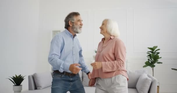 Kaukasiska gamla gråhåriga gifta par dansar hemma och ler. Senior vacker kvinna och stilig man tittar på varandra med kärlek och dans. Manliga och kvinnliga pensionärer lyssnar på musik. — Stockvideo