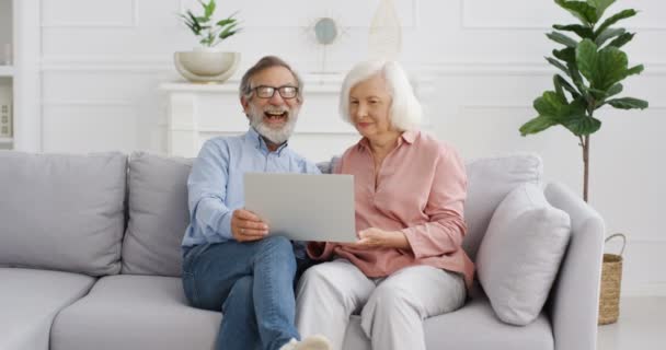 Kaukasisch senior grijs harig gepensioneerd getrouwd stel zittend op de bank en video kijkend op laptop computer. Thuis. Oude gepensioneerden, man en vrouw glimlachend en rustend op de bank. — Stockvideo