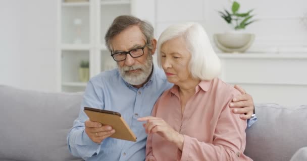 Vit senior gråhårig pensionerad gift par sitter på soffan och tittar på video på tablett enhet. Hemma. Gamla pensionärer, man och kvinna ler och vilar på soffan med gadget dator. — Stockvideo