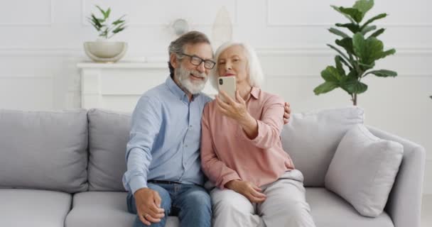 Kaukasische Seniorinnen und Senioren im Ruhestand. Glückliches Paar auf dem Sofa sitzend und lächelnd vor der Handykamera zu Hause posierend. Altes Paar macht Selfie-Fotos mit Smartphone. — Stockvideo