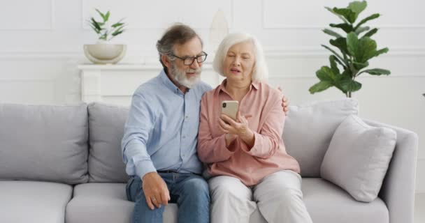 Vieja pareja alegre caucásica sentada en el sofá y viendo algo en la pantalla del teléfono inteligente y hablando. En casa. Hombre y mujer jubilados mayores usando el teléfono móvil, tocando y desplazándose, discutiendo. — Vídeo de stock