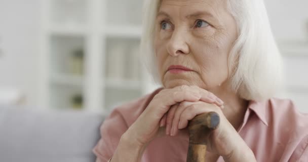 Retrato de la vieja mujer blanca sabia hermosa mirando al lado y pensando con ojos astutos mientras se sienta en el sofá en la sala de estar. Primer plano de la abuela mayor seria con bastón. Casa descansando. — Vídeo de stock