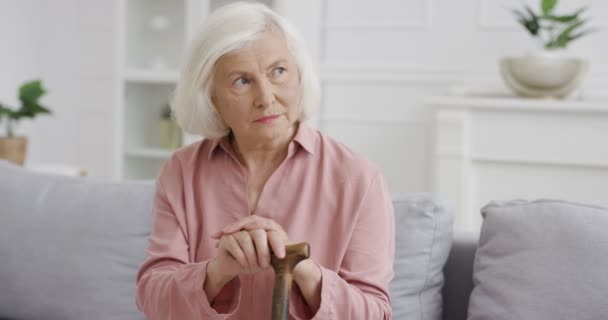 Portrét staré bělošské moudré důchodkyně dívající se stranou a přemýšlející bystrýma očima, zatímco sedí na pohovce s holí v obývacím pokoji Vážná šedovlasá starší babička s holí Domácí odpočinek — Stock video