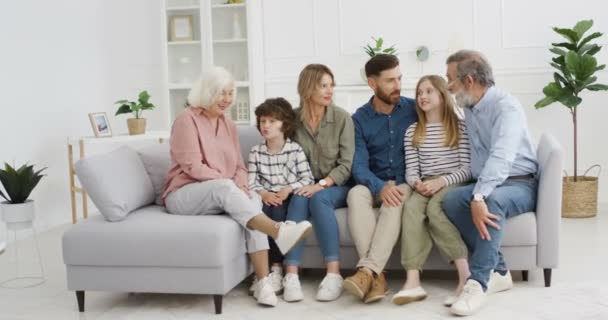 Caucasian happy family with kids sitting on couch in hugs and talking cheerfully. Little boy and girl with parents and grandparents. Three generations. Man and woman with children and grandchildren. — Stock Video