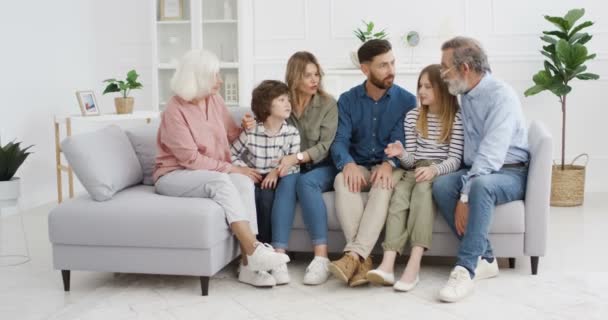 Starsi dziadkowie siedzą na kanapie i radośnie rozmawiają z dziećmi i wnukami. Szczęśliwy mężczyzna i kobieta ze starymi rodzicami i dziećmi na kanapie spędzają razem czas. — Wideo stockowe