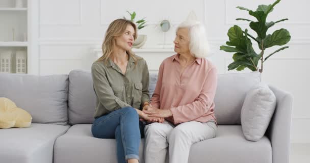 Caucasian beautiful adult woman sitting on couch in cozy living room with senior mother, holding hands and talking with love. Old gray-haired lady with daughter having nice talk and sharing secrets. — Stock Video