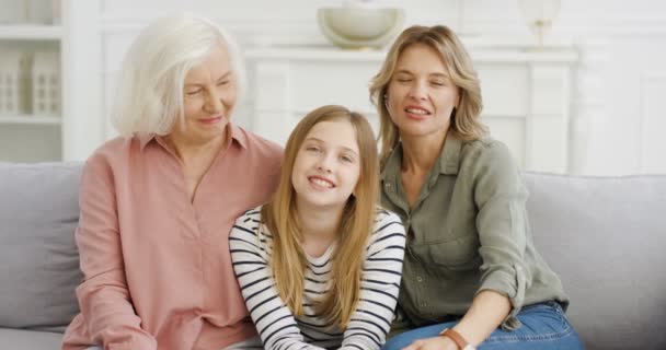 Портрет белой симпатичной девушки-подростка, сидящей на диване с мамой и бабушкой, разговаривающей и машущей руками. Старшая женщина с дочерью и внучкой дома. Поколения женщин. — стоковое видео
