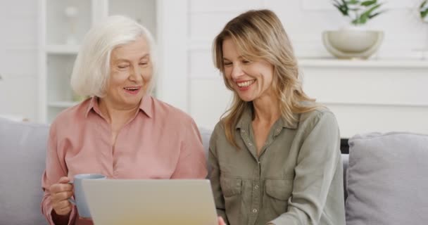 Молода симпатична кавказька жінка сидить на дивані у вітальні з старшою матір'ю і показує щось на ноутбуці. Стара жінка п'є чай і балакає з дорослими доньками.. — стокове відео