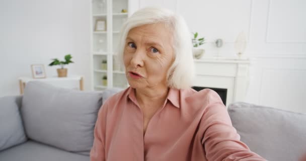 Punto de vista de buena vieja bloguera mujer caucásica con el pelo gris en la sala de estar y hablando con la cámara. Vlogger mujer diciendo algo y explicando. Video blogging. Vlog desde casa. Videochat en webcam. — Vídeo de stock