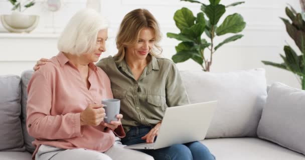 Vacker vit vuxen dotter visar något på laptop till gamla gråhåriga mor wih kopp te. Kvinnor i olika åldrar sitter på soffan och använder dator hemma. Senior kvinna dricker kaffe. — Stockvideo