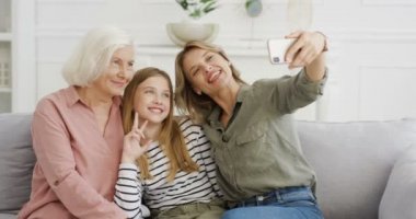 Kafkasyalı genç kız, anne ve büyükannesi ile koltukta oturuyor, akıllı telefon kamerasına kalp işaretleriyle poz veriyor ve selfie çekiyor. Kızı ve torunu olan yaşlı bir kadın evde telefonuyla bekliyor.