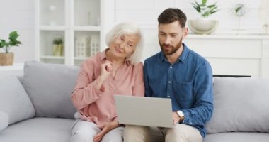 Beyaz, genç, yakışıklı, yetişkin oğlu, yaşlı, gri saçlı annesine rahat oturma odasındaki kanepede dizüstü bilgisayar kullanmayı öğretiyor. Yaşlı kadın bilgisayar kullanıyor ve soru soruyor. Bir adam yazarken ve açıklarken..