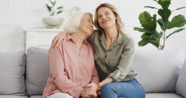 Portrait shot of Caucasian young pretty senior mother and adult young daughter sitting on couch at home and hugging with love. Two females ages and generations. Women embracing, holding hand in hand. — Stock Video