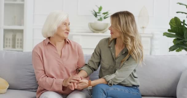 Young Caucasian woman sitting on sofa at home, holding her mother hand and talking together. Gray-haired senior lady with adult daughter having sincere talk. Motherhood love and understanding. — Stock Video