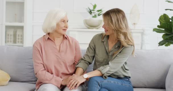 Junge kaukasische Frau, die auf dem Sofa im Wohnzimmer sitzt, ihre Mutter an der Hand hält und miteinander redet. Grauhaarige Seniorin mit erwachsener Tochter, die aufrichtige Gespräche führt. Mutterliebe und Verständnis. — Stockvideo