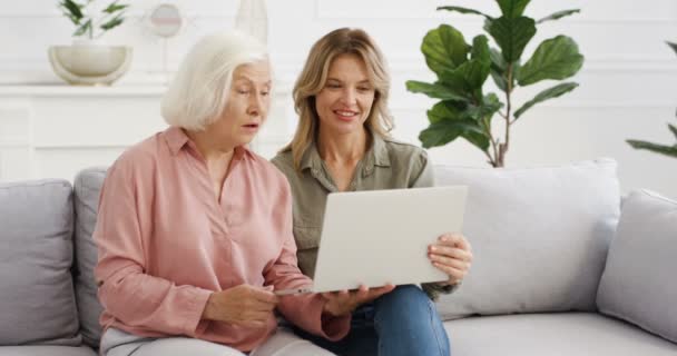 漂亮的白人成年女儿在笔记本电脑上给白发老太婆展示一些东西，并讨论这个问题。不同年龄段的妇女坐在沙发上，在家里用电脑。老太婆说话. — 图库视频影像