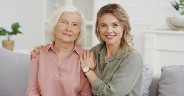 Portrait of beautiful Caucasian young woman hugging senior mother pensioner with gray hair while sitting on couch at home. Adult pretty daughter and old mom embracing and smiling to camera. Indoor. — Stock Video