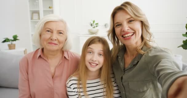 Beautiful Caucasian three female generations. Pretty cheerful woman with teen small daughter and senior mother talking to camera and videochatting indoor. POV. Videochat on webcam with grandmother. — Stock Video