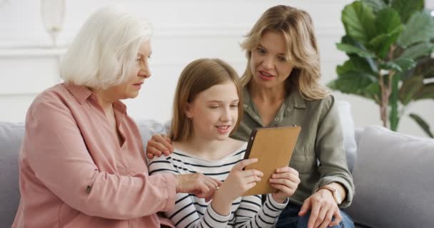 Roztomilý kavkazský dospívající dívka pomocí tabletu zařízení a sledování něco na obrazovce s matkou a babičkou na gauči v obývacím pokoji. Starší žena s dcerou a vnučkou klepání na gadget. — Stock video