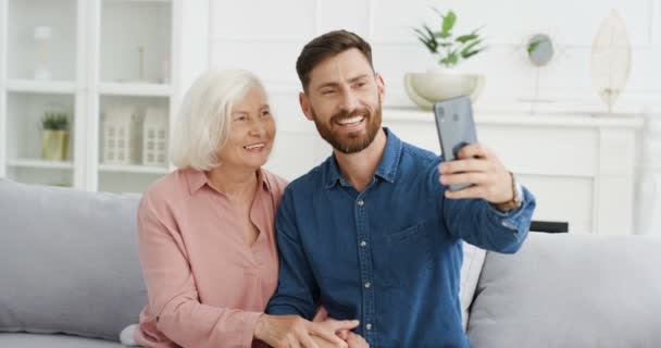 Pria tampan kaukasia mengambil foto selfie dengan kamera smartphone dengan wanita berambut abu-abu senior di sofa di rumah. Anak laki-laki tersenyum dewasa berpose ke telepon dengan ibu tua di ruang tamu. — Stok Video
