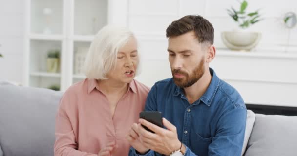 Blanc jeune beau fils adulte enseignant à sa vieille mère aux cheveux gris d'utiliser un smartphone sur le canapé dans le salon confortable. Femme âgée posant des questions et regardant l'homme tapoter et défiler au téléphone. — Video