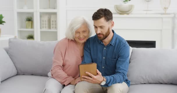 Biały, młody, przystojny syn uczy swoją starą, siwą matkę używać tabletu na kanapie w przytulnym salonie. Starsza kobieta za pomocą gadżetu komputerowego i zadaje pytania, gdy mężczyzna stuka. — Wideo stockowe