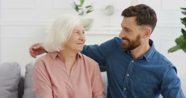 Portret van een blanke jonge knappe man die glimlacht en zijn oude grijze moeder knuffelt terwijl ze op de bank zitten in de woonkamer. Oudere moeder in knuffels met volwassen zoon. Binnenshuis. — Stockvideo