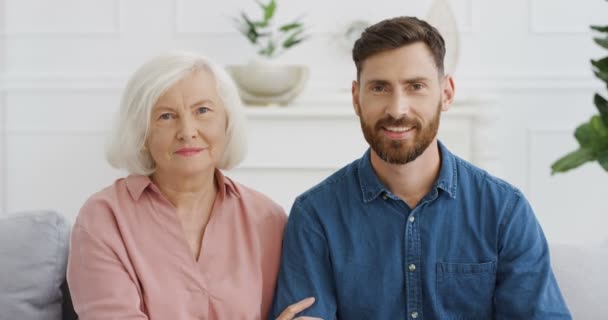 Portrait shot of Caucasian old gray-haired woman hugging her adult son and smiling to camera while they sitting on sofa in living room. Handsome man embracing senior smiled mother at home. Indoor. — Stock Video